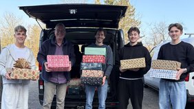Vier männliche Personen stehen vor dem geöffneten Kofferraum eines Transpoeters. Sie halten schön verpackte Geschenke in ihren Händen. Sie lächeln in die Kamera.