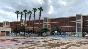 Ein großes langgestrecktes Schulgebäude aus roten Ziegeln. Davor stehen mehrere hohe Palmen.